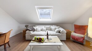 Une pièce de style loft avec un plafond en pente, un canapé blanc, une table à manger avec une bouteille et des verres, une chaise en osier et une chaise rouge.