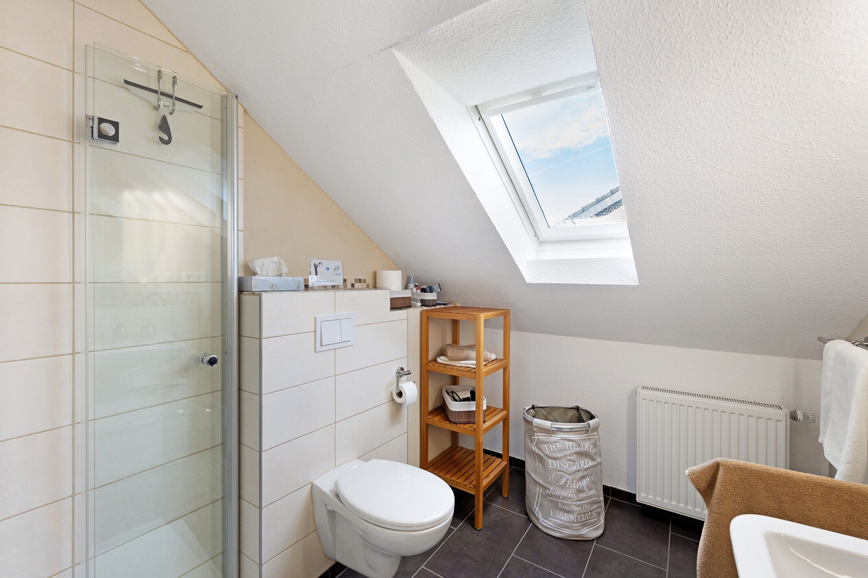 Une salle de bain moderne avec une douche à parois de verre, des toilettes blanches, une étagère en bois et un puits de lumière.