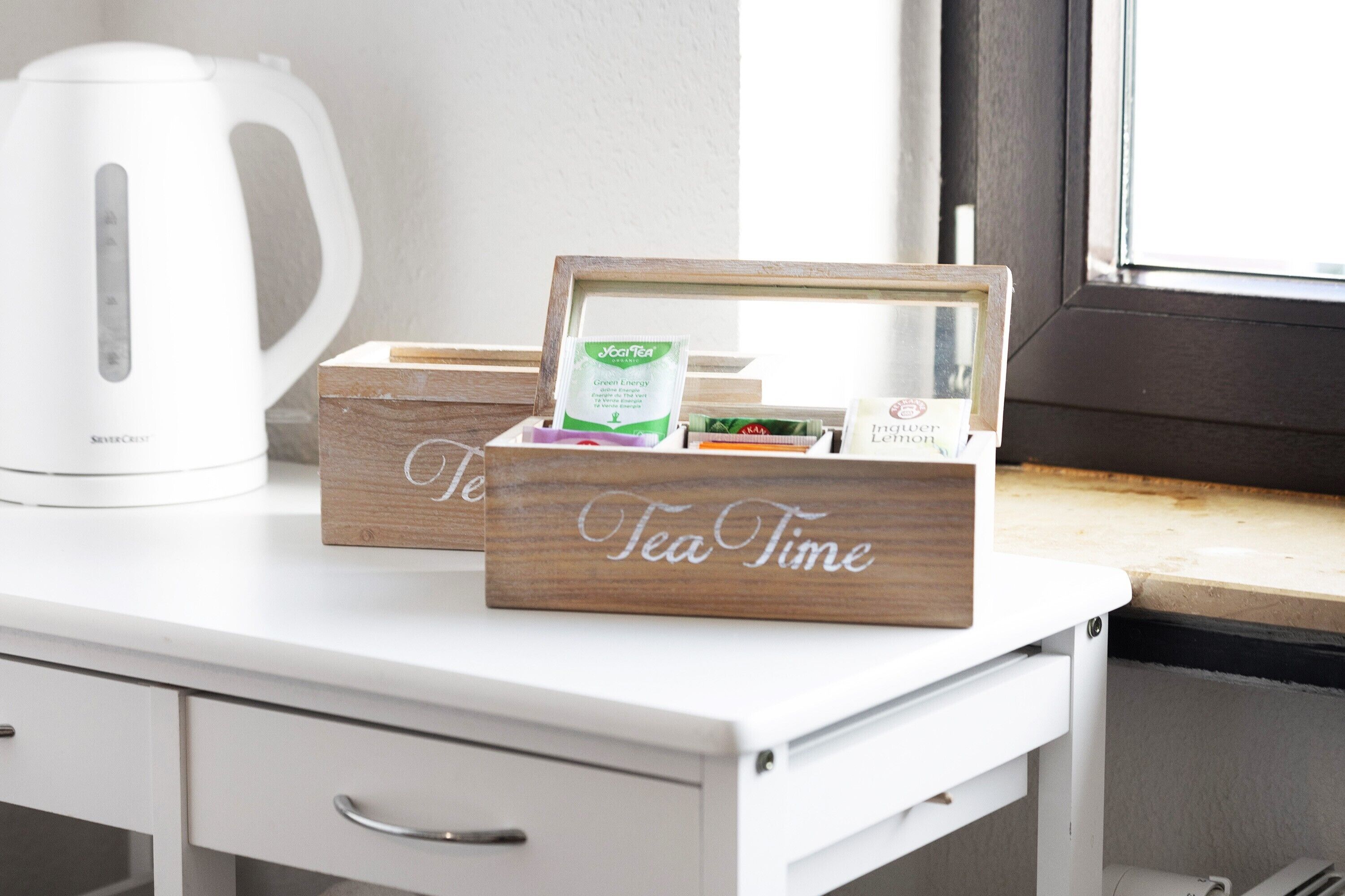 Una taula blanca amb una olla elèctrica blanca, dues caixes de fusta amb la inscripció 'Tea Time' i un objecte emmarcat amb text.