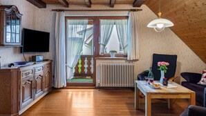 Una sala de estar acogedora con techo de madera, un televisor de pantalla plana, una mesa de comedor con un jarrón de flores y una ventana con cortinas.