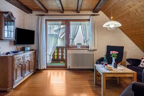 Una sala de estar acogedora con techo de madera, un televisor de pantalla plana, una mesa de comedor con un jarrón de flores y una ventana con cortinas.