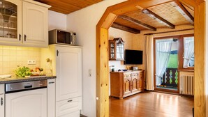 Una cocina con armarios blancos, microondas y lavavajillas. Una puerta de madera da a una sala de estar con televisor y ventana.