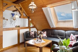 Ein gemütliches Zimmer mit Holzbalkendecke, einem schwarzen Ledersofa, einem Holztisch mit Flasche und Gläsern sowie einem großen Fenster mit Vorhängen.