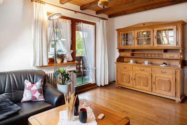 Un salon avec un canapé en cuir, un meuble en bois, une table ornée d’un vase et de verres, et une fenêtre avec des rideaux.