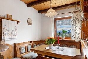 Un coin repas avec une table en bois, des bancs, un vase de fleurs, une horloge et une fenêtre donnant sur un paysage.