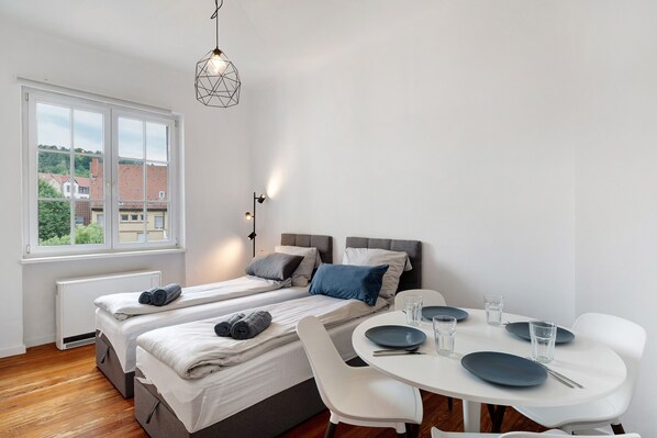A bedroom with two beds, a dining table, and a view of buildings outside.