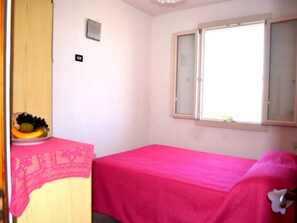 A small room with a single bed, a window with a view, a wall-mounted light switch, and a fruit bowl on a table.