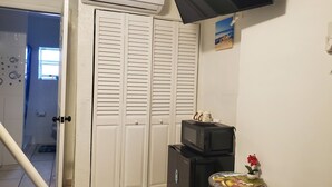 Una habitación con un armario blanco cerrado, un refrigerador negro, un microondas encima, una mesita con un plato decorativo y un televisor montado en la pared.