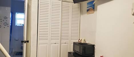 Una habitación con un armario blanco cerrado, un refrigerador negro, un microondas encima, una mesita con un plato decorativo y un televisor montado en la pared.