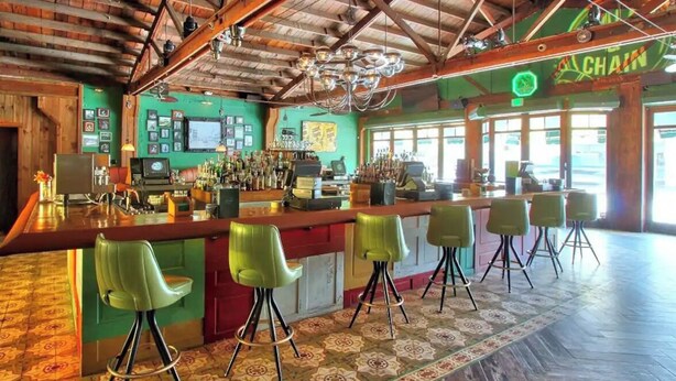Un bar con techo de madera rústica y paredes verdes, que presenta una barra con taburetes y un letrero de neón.