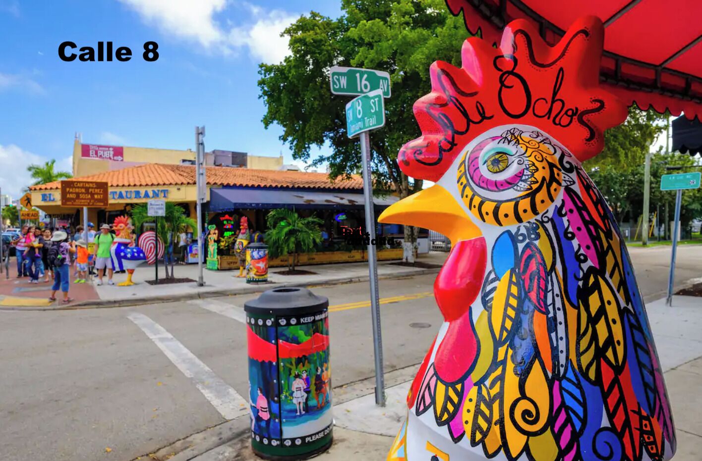 A colorful rooster statue on a street corner with a restaurant named 'El Pub' in the background.