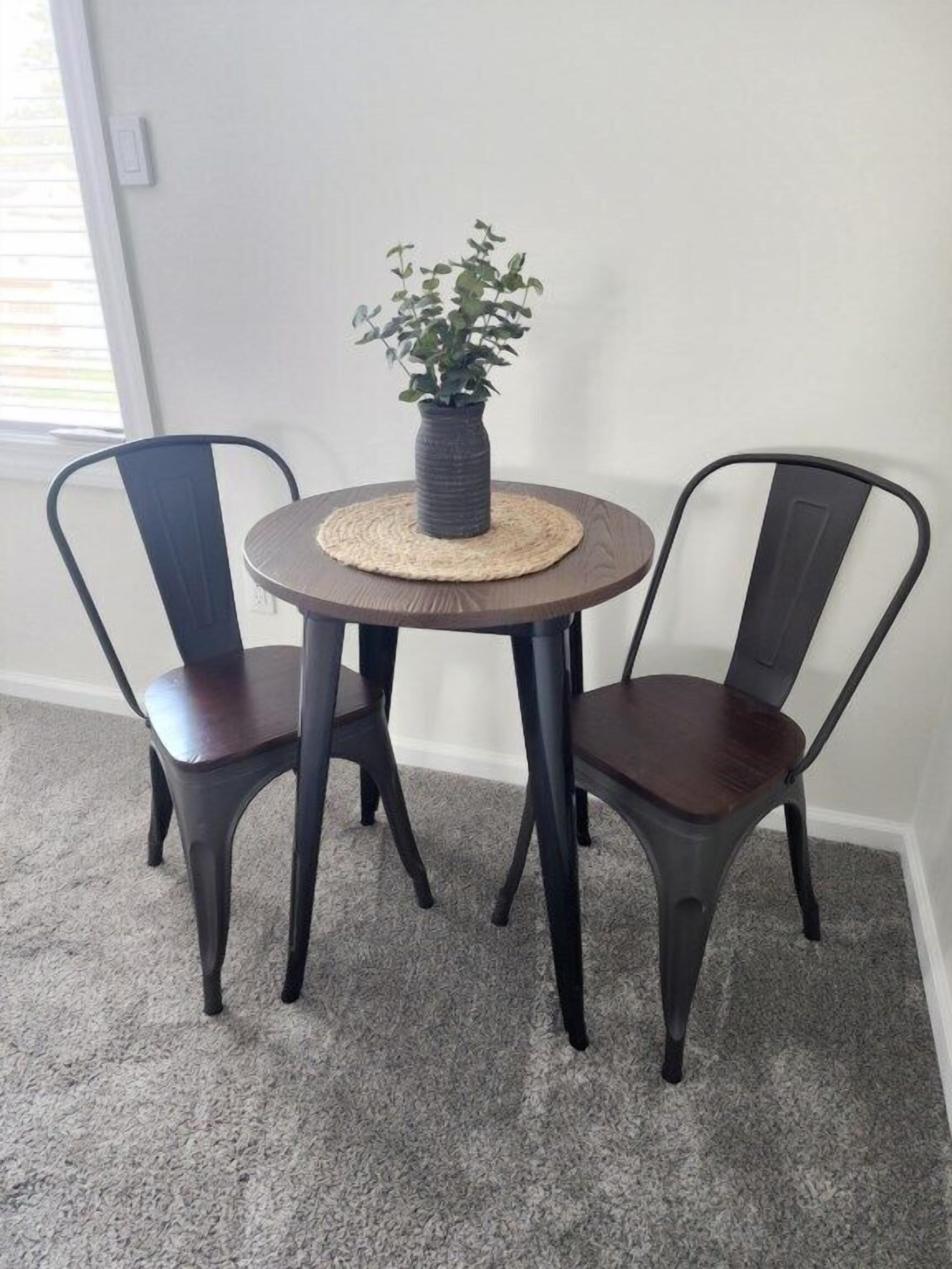 A round table with a woven placemat and a potted plant in the center, flanked by two dark metal chairs.