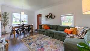 Un salon avec un canapé vert, une table à manger en bois avec des chaises, un tapis coloré et une grande fenêtre donnant sur l’extérieur.