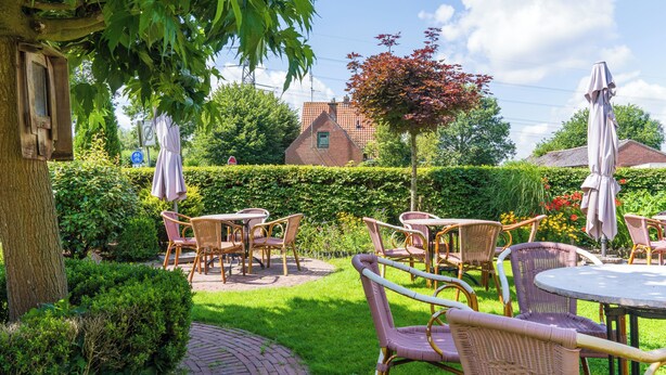 Eine Terrasse mit Korbmöbeln und Sonnenschirmen, umgeben von Grünpflanzen und einem Backsteinhaus im Hintergrund.