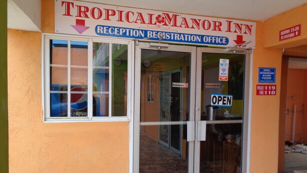 Entrada a la recepción y oficina de registro del Tropical Manor Inn, con puertas de vidrio, un letrero que indica la disponibilidad de Wi-Fi y números de contacto de emergencia.
