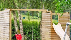 Une structure de jeux en bois, dotée d’un toboggan et de balançoires, entourée de verdure.