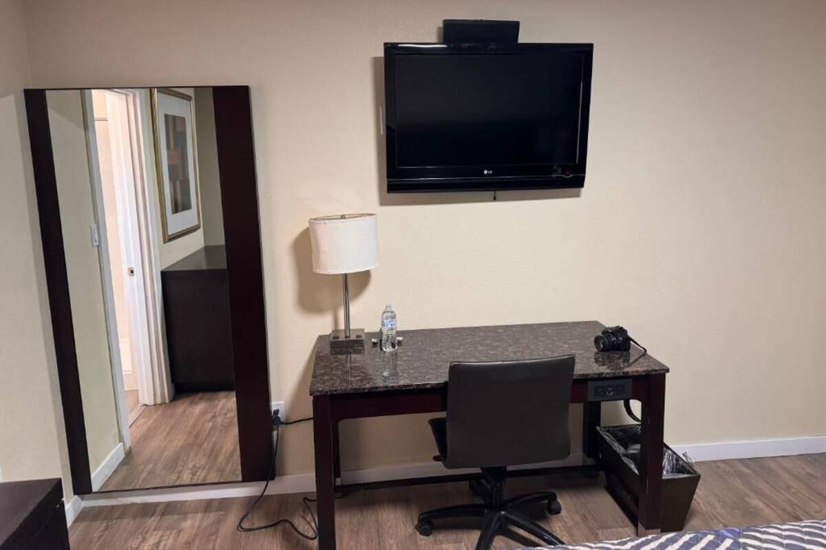 A hotel room with a desk, chair, lamp, and a flat-screen TV mounted on the wall.