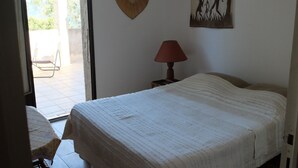 A bedroom with a bed, a nightstand with a lamp, a wall hanging, and a view of the outside. - Loue Maison de Famille Dans le Golf de Lava à Proximité de la mer (Villanova)