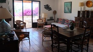 A spacious living area with a dining table, chairs, a sofa, and a large window offering a view of pink flowers. - Loue Maison de Famille Dans le Golf de Lava à Proximité de la mer (Villanova)