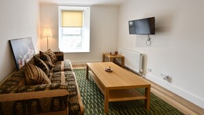 A living room with a sofa, a wooden table, a TV mounted on the wall, and a window with a blind. - Troon Portland Apt B (Troon)