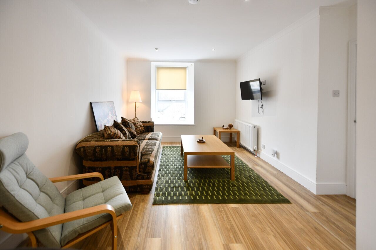 A living room with a sofa, armchair, and coffee table. There is a window with blinds, a radiator, and a TV mounted on the wall.