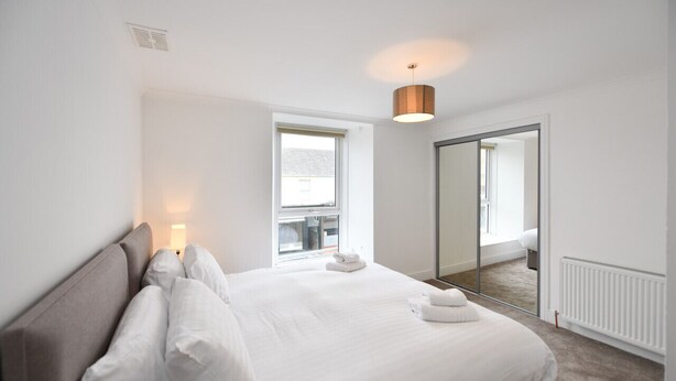 A modern hotel room with a large bed, a bedside lamp, a window with a view, and a mirror on the wall. - Troon Portland Apt B (Troon)