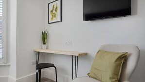 Ein Zimmer mit weißer Wand, einem Flachbildfernseher über einem Holztisch, einem schwarzen Stuhl, einem weißen Sessel mit grünem Kissen und einem Blumentopf auf dem Tisch.