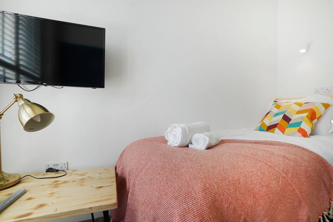Habitación de hotel con cama, televisión montada en la pared, escritorio con lámpara y un cojín colorido.