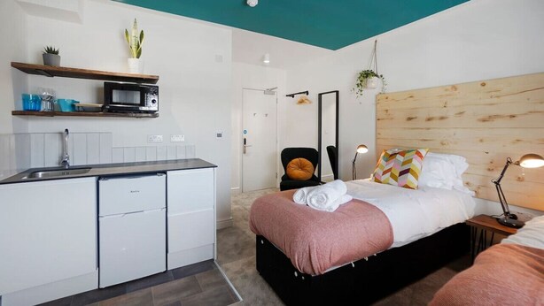 Quarto de hotel moderno com cozinha compacta, cama com cabeceira de madeira e banheiro com porta branca.