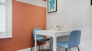 Quarto com uma mesa branca e duas cadeiras azuis, um quadro abstrato emoldurado na parede e uma pequena planta sobre a mesa.