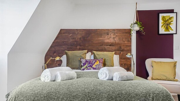 A bedroom with a wooden headboard, a green textured bedspread, two white towels, a purple floral pillow, and a yellow pillow. - Eco Aparthotel - Heights Studio (Folkestone)
