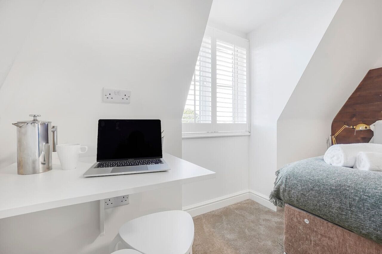 Um quarto pequeno e branco, com uma escrivaninha, um laptop, uma cama e uma janela com persianas.