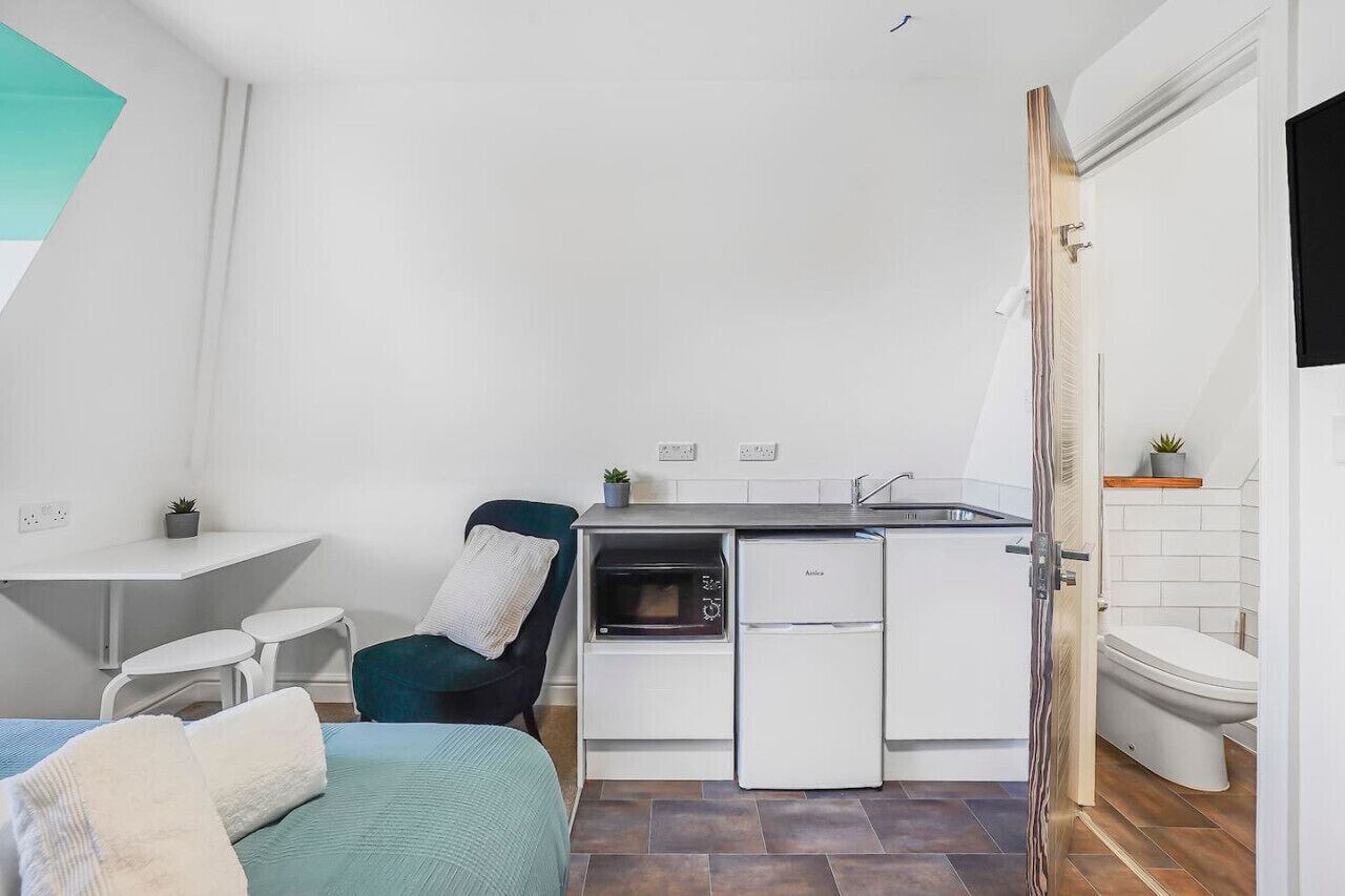 Een compacte keuken met een magnetron, oven en koelkast, een klein eetgedeelte met een tafel en stoelen, en een enkel bed met kussens.