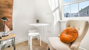 A corner room with a white desk, a chair with an orange cushion, a small potted plant, and a window with a view of buildings. - Eco Aparthotel - Heights Studio (Folkestone)