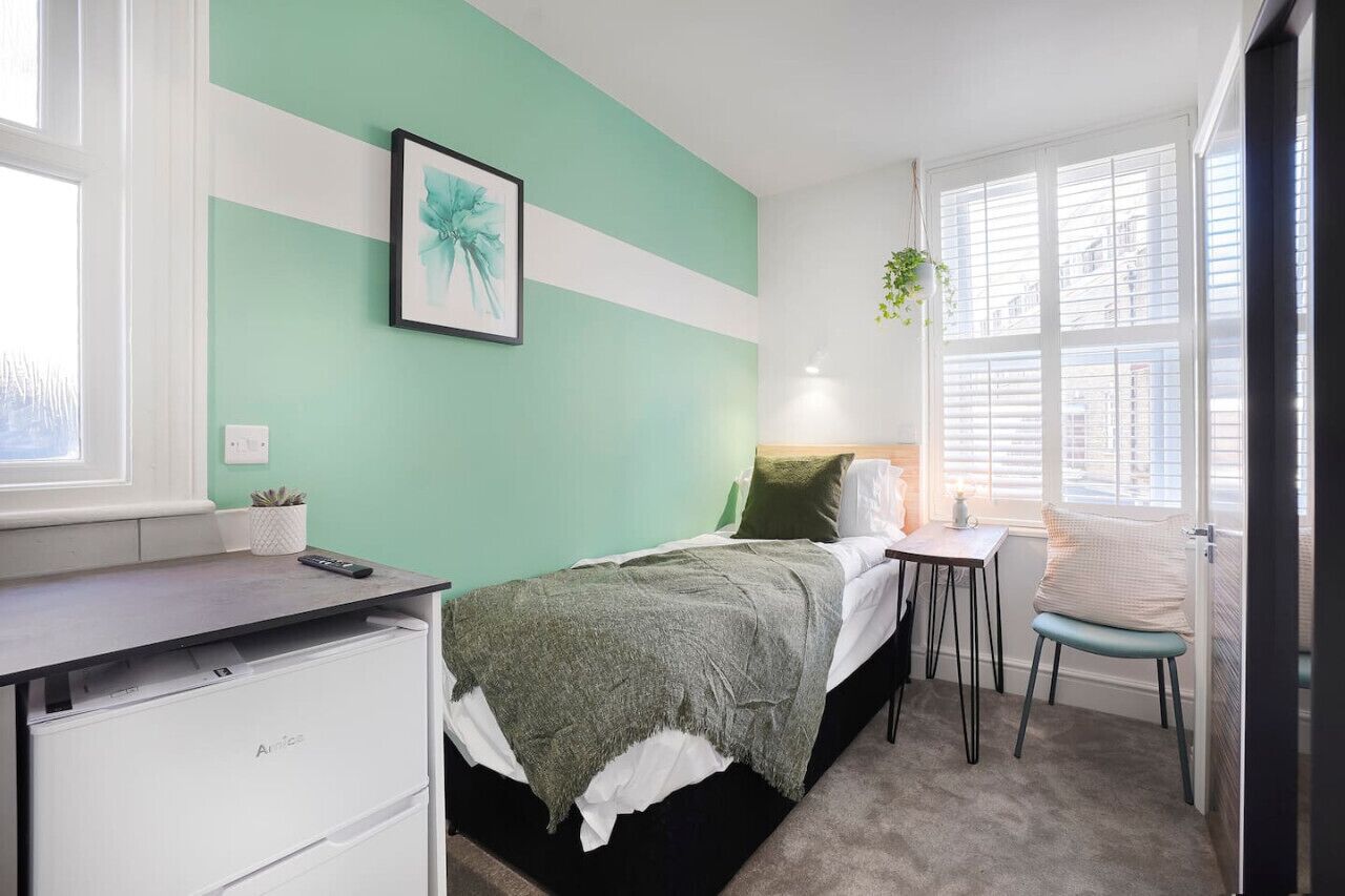 Un dormitorio con cama, escritorio, silla, ventana con persianas y una planta.