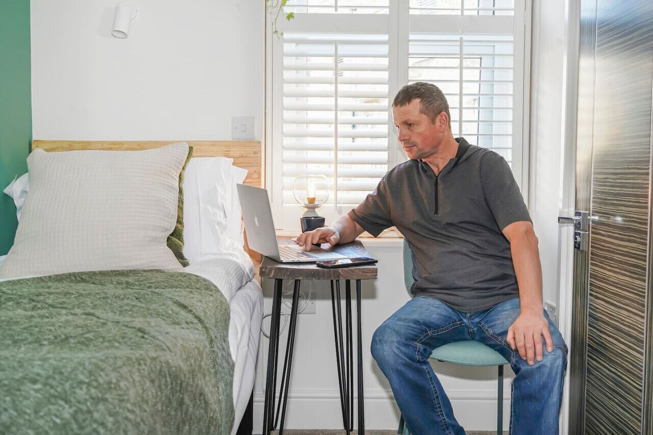 Ein Mann sitzt auf einem Hocker an einem kleinen Tisch mit einem Laptop in einem Zimmer, das ein Bett, ein Fenster mit Jalousien und einen Kühlschrank enthält.