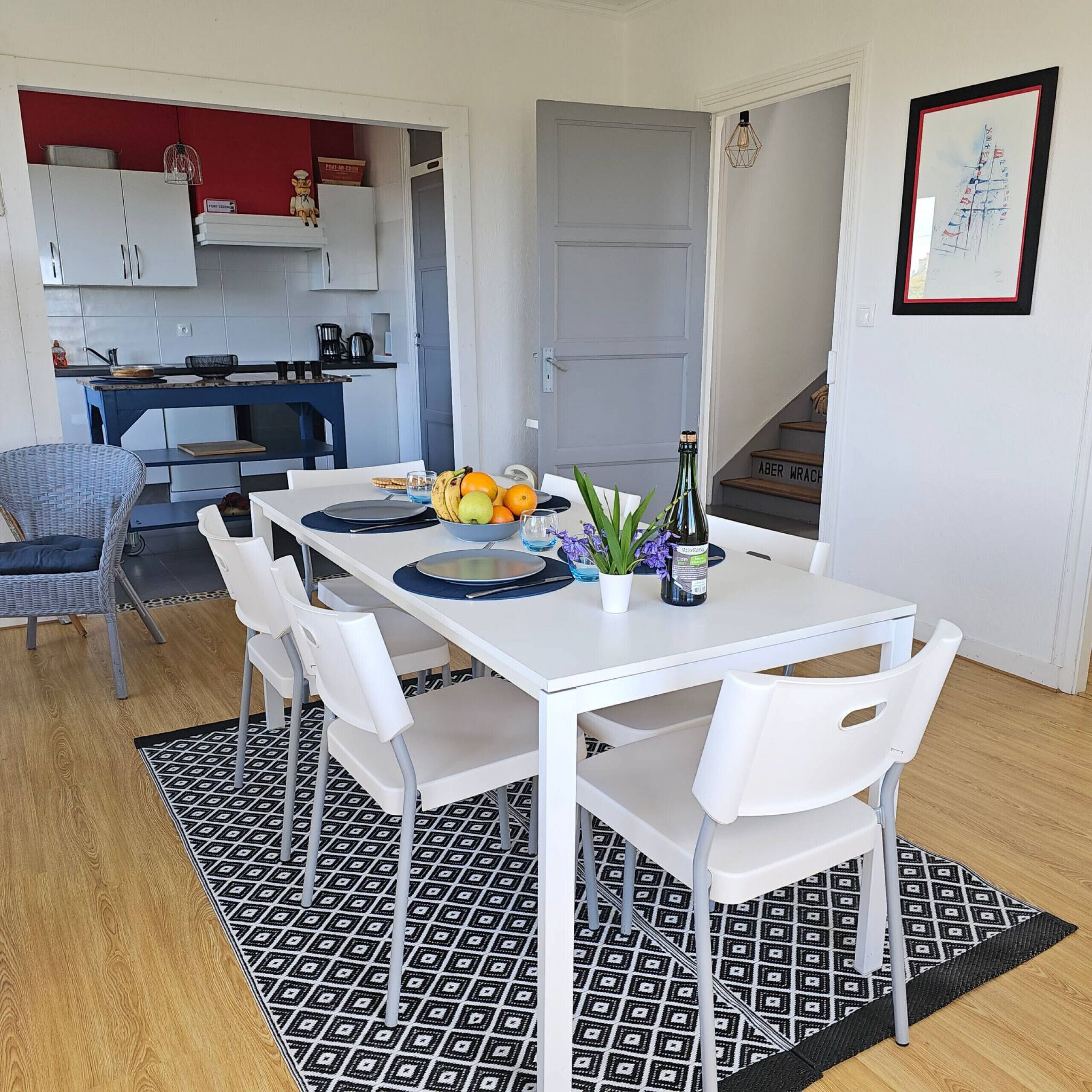 Ein Essbereich mit einem weißen Tisch, gedeckt für vier Personen, umgeben von weißen Stühlen. Auf dem Tisch befinden sich eine Obstschale, eine Weinflasche und ein Blumentopf. Der Raum verfügt über einen gemusterten Teppich, eine Küche im Hintergrund und eine Treppe, die zu einer anderen Etage führt.