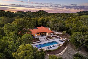 Une maison avec une piscine, entourée d’une végétation luxuriante et avec des collines en arrière-plan.