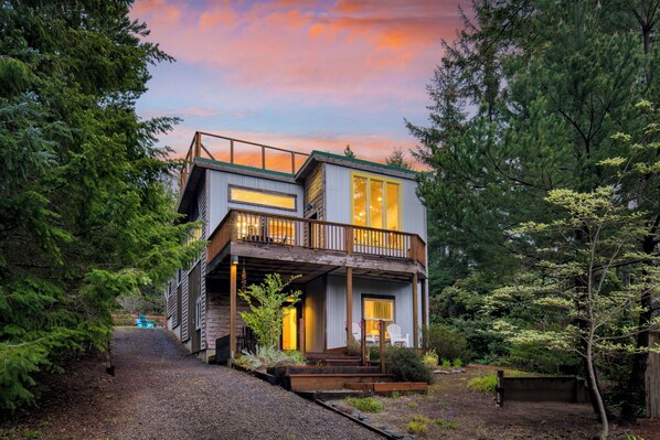 A two-story wooden house with a balcony, surrounded by trees and a gravel driveway.