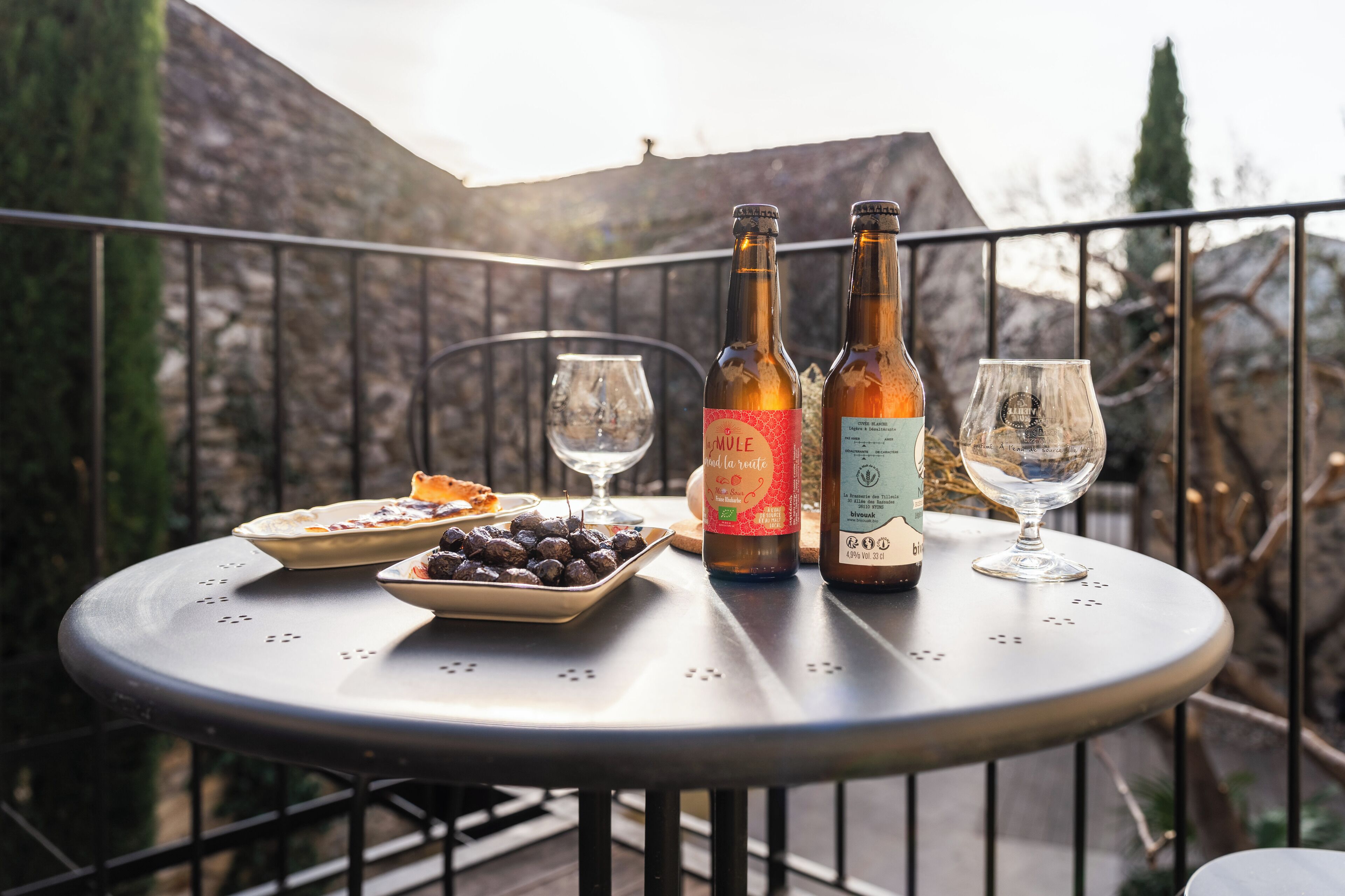 A round metal table with two bottles of wine, two wine glasses, a plate of food, and a bowl of olives on a balcony.