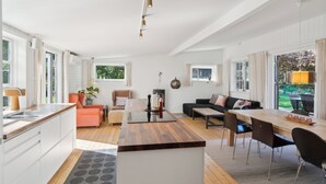 A modern kitchen with a wooden island, a dining area with a long table and chairs, a sofa, and a view of the garden. - Spacious summer house with pool. (Vejby)