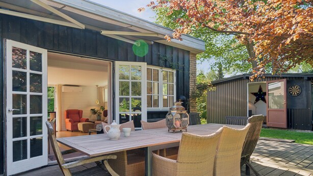 An outdoor deck with a table and wicker chairs, a sliding glass door, and a dartboard. - Spacious summer house with pool. (Vejby)