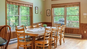 A dining room with a wooden table and chairs, large windows with wooden frames, and a view of the outdoors.