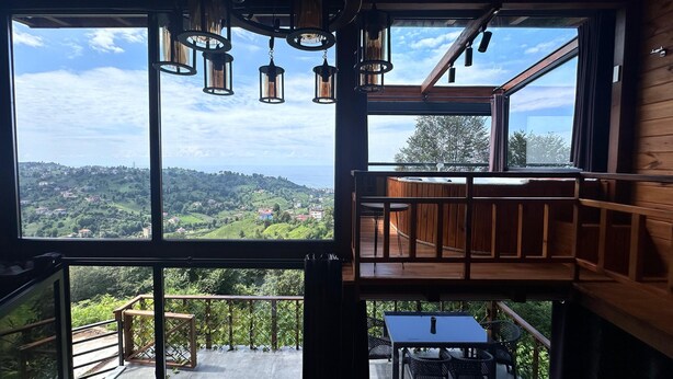 Une pièce dotée de grandes fenêtres donnant sur un paysage vallonné. On y trouve une terrasse en bois avec une table et des chaises.