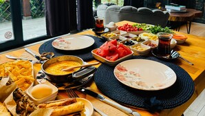 Une table à manger dressée avec une variété de plats, notamment du pain, de la soupe et des fruits, dans une pièce dotée d’une cheminée en pierre et offrant une vue sur la verdure environnante.