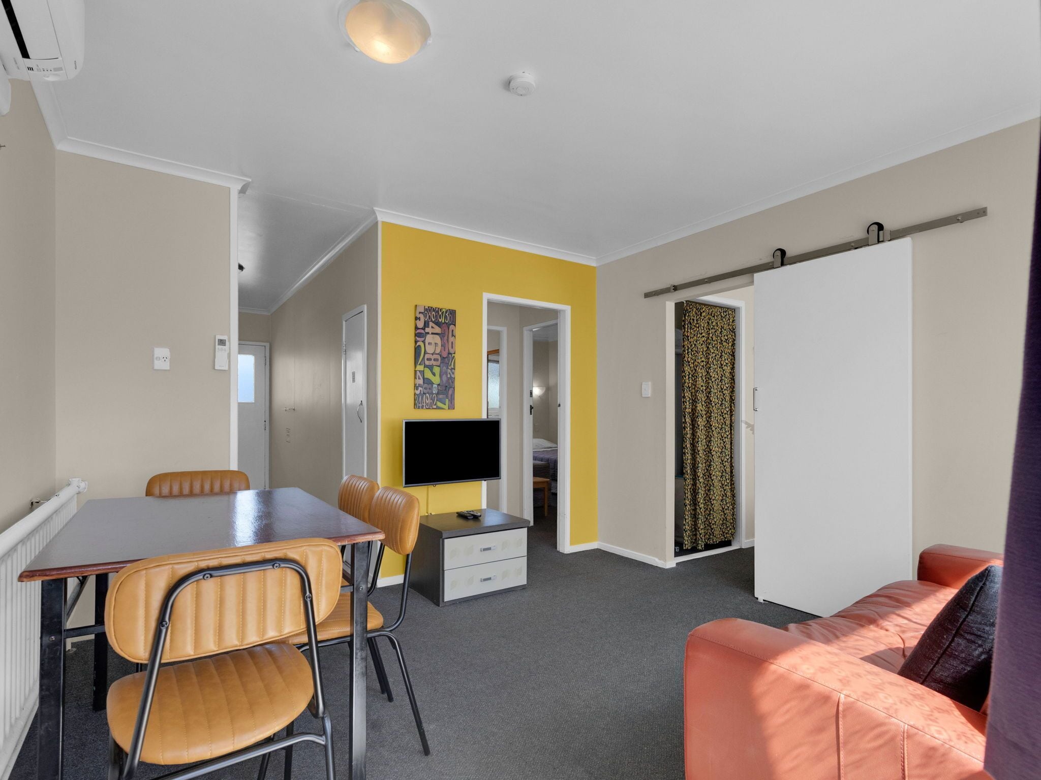 Een kleine kamer met een eettafel, stoelen, een bank, een televisie en een deur naar een andere kamer.