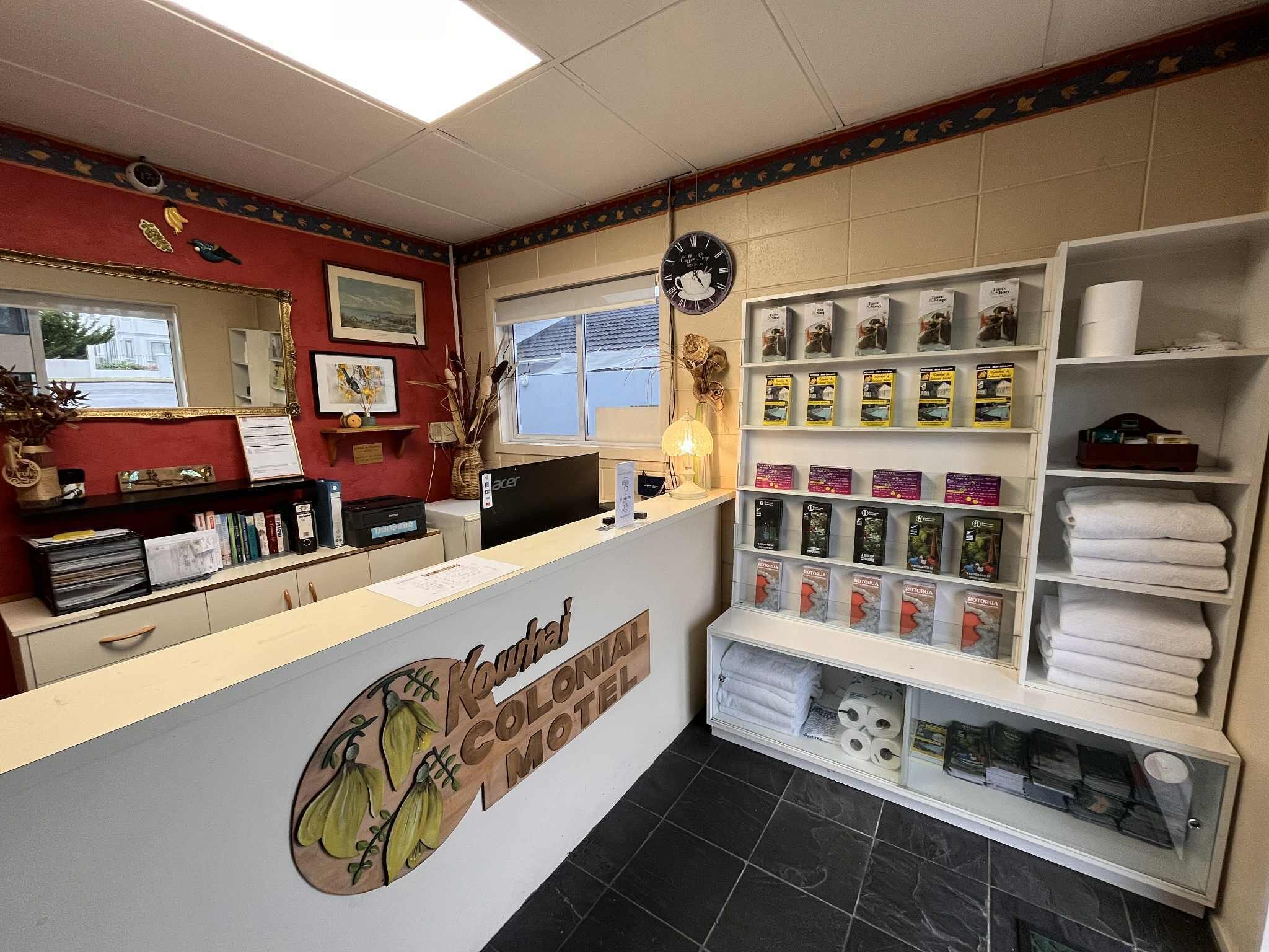 Un mostrador de recepción con un cartel que dice 'Kuwana Colonial Motel'. Hay estantes con libros y artículos de aseo, un reloj y una ventana con vistas al exterior.