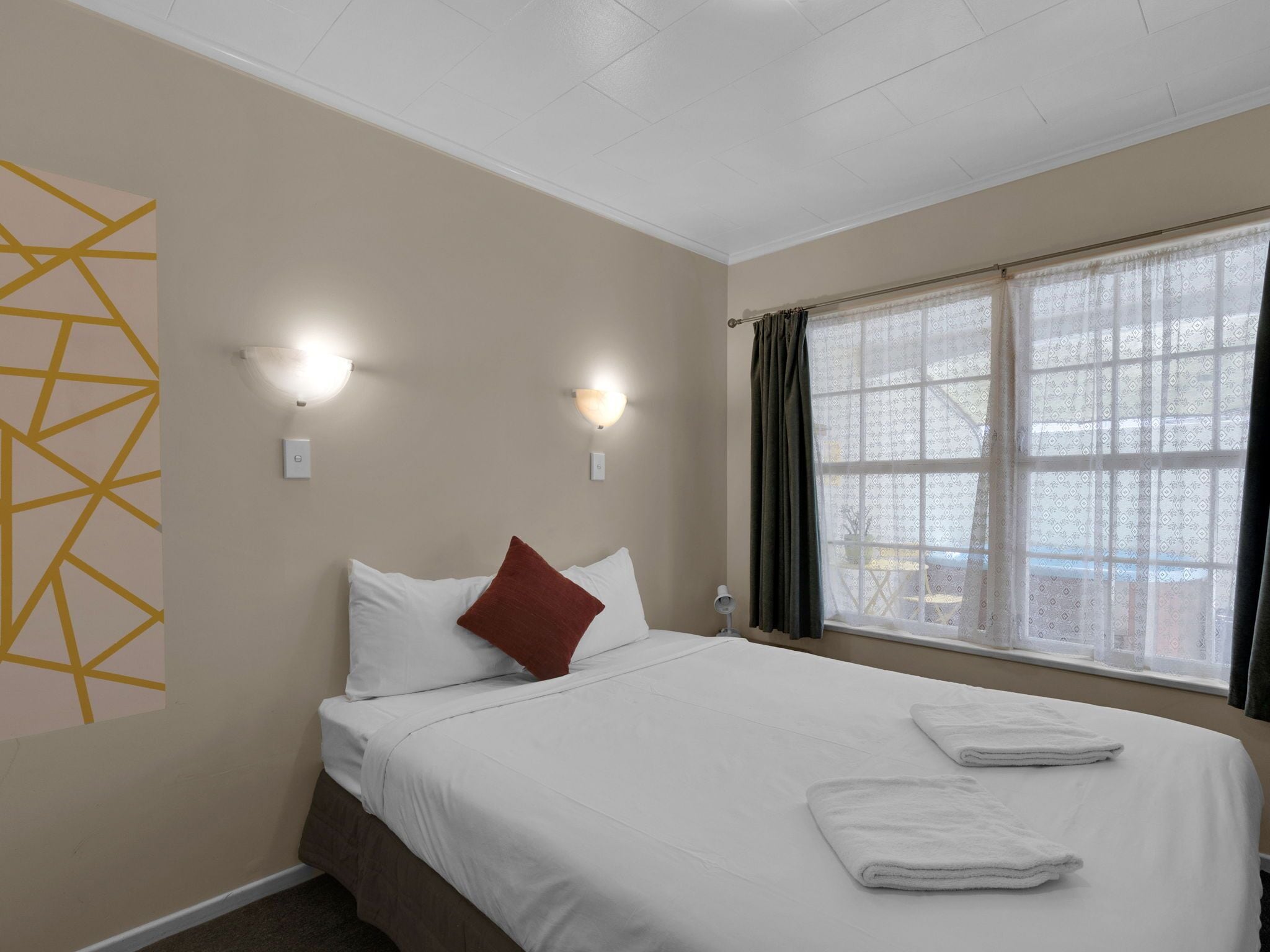 A hotel room with a bed, two wall-mounted lights, a window with curtains, and a geometric wall decoration.