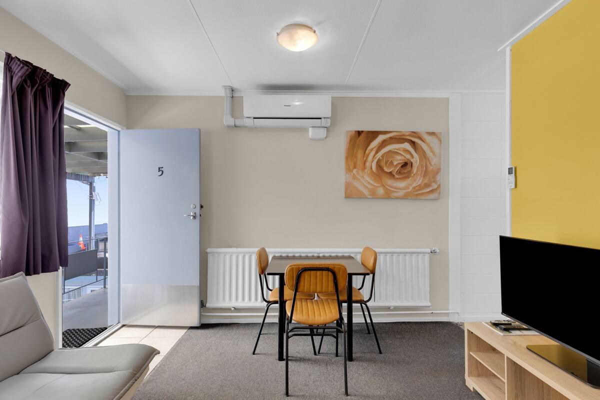 A modern living room with a sofa, a dining table with chairs, a flat-screen TV, a radiator, and a door marked with the number 5.