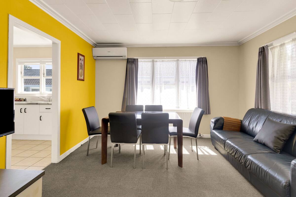A room with a dining table, chairs, a sofa, a TV, and an air conditioning unit.